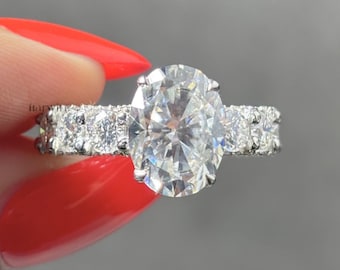 May include: A silver-coloured ring featuring a large, oval-cut centre stone held by four prongs. The band is encrusted with a series of smaller, round-cut stones extending along both sides. The metal has a polished, reflective finish throughout.