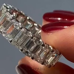 May include: A close-up of a silver eternity ring with multiple emerald-cut diamonds. The ring is held between fingers with red nail polish. The diamonds sparkle, reflecting light.