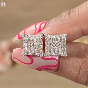 May include: Gold square earrings, each adorned with a multitude of small, clear gemstones. The earrings feature a rectangular design, with a border of smaller stones encircling a central cluster of larger, rectangular-cut gems. The earrings are held by a person's fingers.
