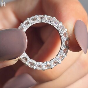Könnte beinhalten: Ein silberfarbener Eternity-Ring mit einer durchgehenden Reihe runder, klarer, facettierter Steine in einer Metallfassung. Der Ring hat eine glänzende Oberfläche, wobei die Steine den gesamten Umfang des kreisförmigen Bandes umschließen.