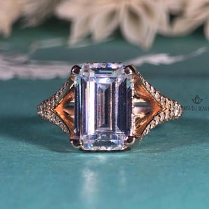 Könnte beinhalten: Ein Ring aus Roségold mit einem großen, im Smaragdschliff geschliffenen Mittelstein, der von vier Krappen gehalten wird. Die Ringschiene ist gespalten und an den Seiten mit kleinen Pavé-gefassten Steinen besetzt. Das Metall glänzt warm.