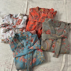May include: Four floral patterned robes in different colors, including teal, orange, white, and green. The robes are folded and arranged on a textured beige surface.