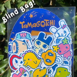 Può includere: Un sacchetto a sorpresa di adesivi Tamagotchi con vari personaggi in diversi colori e pose. Il sacchetto è blu con stelle bianche e il testo "Tamagotchi" in bianco.