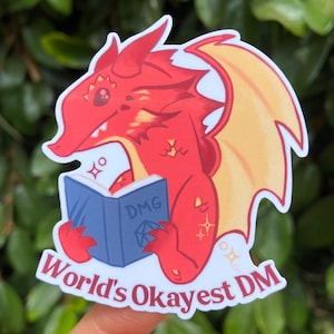 May include: A red dragon sticker with yellow wings, holding a blue book labeled "DMG." The sticker reads "World's Okayest DM" in white text. The dragon has a friendly expression and is surrounded by decorative sparkles.