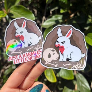 Peut inclure: Deux autocollants découpés représentant un lapin blanc avec des marques rouges sur le visage et la poitrine, assis dans une grotte brune à côté d'un crâne humain et d'os. L'autocollant de gauche a une finition holographique et le texte JUST A HARMLESS LITTLE BUNNY.