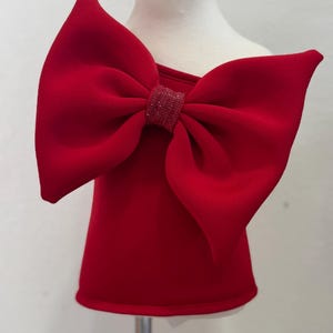May include: A sleeveless red top featuring a large, structured bow on the front shoulder. The center of the bow is embellished with a band of small, sparkling red crystals. The garment is displayed on a mannequin, showcasing a solid red color.