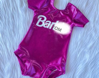 May include: A short-sleeved leotard in a vibrant, metallic fuchsia fabric. The front of the garment features the white text "Barbie" with the word "CUSTOM" printed in smaller black letters layered over the white text on the chest area.
