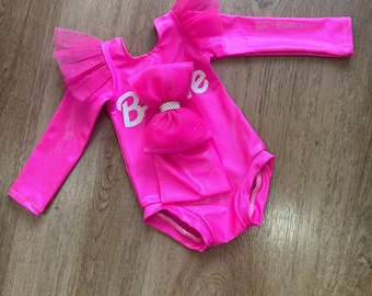May include: A bright hot pink long-sleeve leotard featuring ruffled tulle shoulder accents and a large central bow with a rhinestone center. The garment displays the white text Barbie across the chest, constructed from a smooth, stretchy fabric.