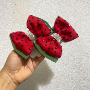 以下が含まれることがあります： スイカをテーマにしたヘアリボン。赤い生地に黒いビーズの種と緑色の皮が付いています。キラキラ光るラインストーンバンドがリボンを飾っています。リボンは、テクスチャ加工された白い背景に対して、手で持たれています。