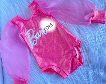 May include: A bright pink glittery leotard featuring long sheer puff sleeves and a round neckline. The front of the garment displays the word "Barbie" in a light blue script font with the word "CUSTOM" printed in white letters above it.