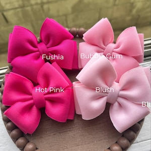 May include: A collection of four fabric hair bows in shades of pink, including fuchsia, bubble gum, hot pink, and blush. Each bow is labeled with its color name. The bows are arranged on a wooden tray with a beaded edge.