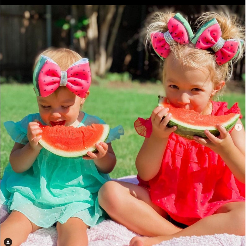 以下が含まれることがあります： スイカをテーマにしたヘアリボンとドレスを着た2人の幼い子供たちが、スイカのスライスを楽しんでいます。リボンはピンク色で、黒い種子のディテールとラインストーンのアクセントが付いています。一人の子供はターコイズのドレスを着ており、もう一人は赤いドレスを着ています。