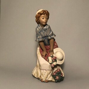 Puede incluir: Figura de cerámica de una mujer con un vestido blanco largo con un delantal rojo y una camisa azul. Sostiene un sombrero blanco y un ramo de rosas. La figura tiene cabello castaño y está mirando hacia abajo.