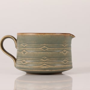 May include: A green ceramic creamer with a brown rim and a geometric pattern. The creamer has a handle and a spout.