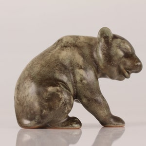 May include: A ceramic bear figurine in a green and brown glaze. The bear is sitting on its haunches with its head tilted down.