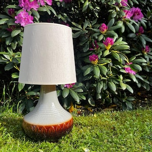 Könnte beinhalten: Eine Keramik-Tischlampe mit beigem Stoffschirm. Der Lampenfuß ist eine Kombination aus Grau, Weiß und Braun mit einem strukturierten Design. Die Lampe steht auf einer grasbewachsenen Oberfläche mit einem Hintergrund aus grünem Laub und rosa Blüten.