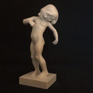 Puede incluir: Una figurita de porcelana blanca de una niña de pie sobre una base cuadrada. La niña está desnuda y tiene el pelo recogido hacia atrás. Está mirando hacia abajo y su brazo derecho está doblado por el codo.