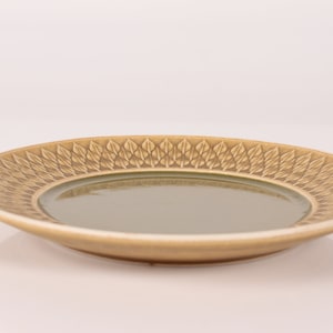 Op de afbeelding: Een lichtbruin keramiek bord met een groene glazuur en een decoratief patroon van gestileerde bladeren rond de rand.