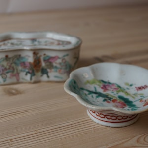 Könnte beinhalten: Zwei kleine, weiße Porzellanschalen mit gewellten Rändern. Beide Schalen sind mit bunten floralen und figürlichen Designs verziert.