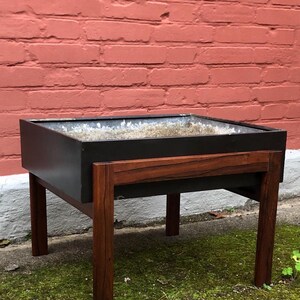 Puede incluir: Una jardinera de mediados de siglo con un contenedor rectangular oscuro y un marco de madera contrastante. La jardinera tiene un contenedor oscuro y está sostenida por cuatro patas de madera. La madera tiene un rico tono marrón rojizo.