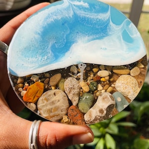 May include: Round resin coaster with a blue and white wave design and a layer of colorful and brown stones at the bottom.