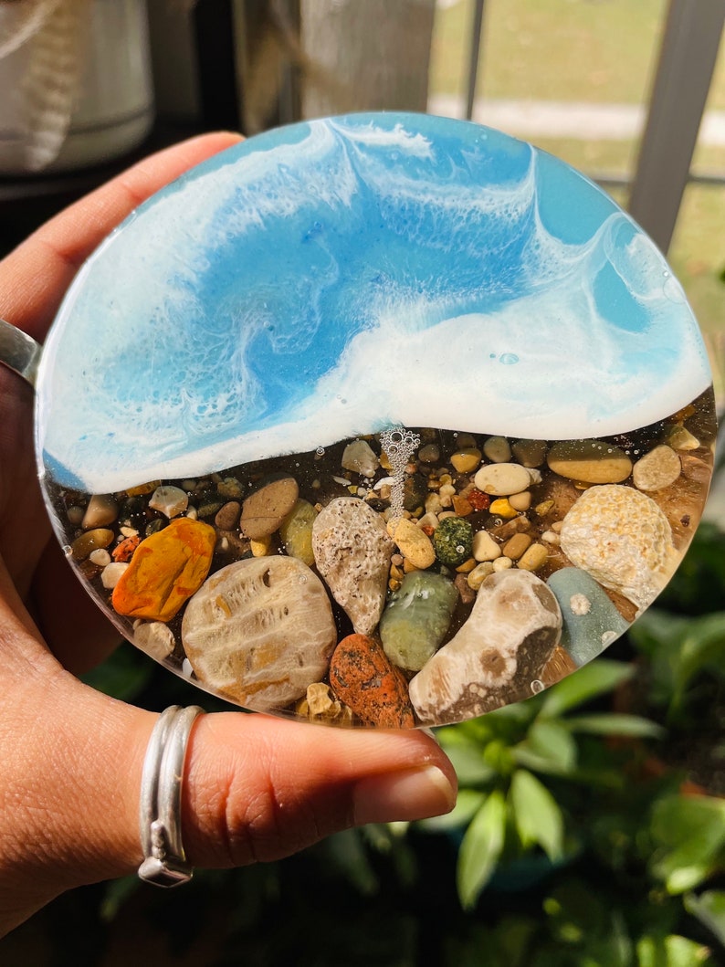May include: Round resin coaster with a blue and white wave design and a layer of colorful and brown stones at the bottom.