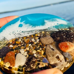 May include: A round, clear resin coaster with a blue and white wave design. The coaster is filled with small pebbles and seashells, including a large, light brown seashell.