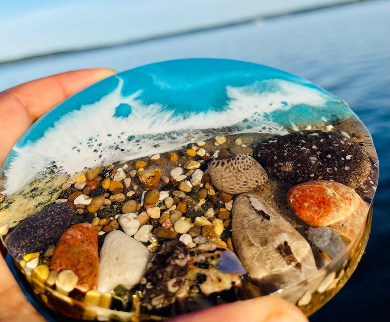 May include: A round, clear resin coaster with a blue and white wave design. The coaster is filled with small pebbles and seashells, including a large, light brown seashell.