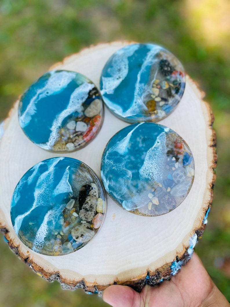 May include: Four round resin coasters with a beach scene. The coasters feature blue water, white foam, and brown sand with small shells and pebbles.