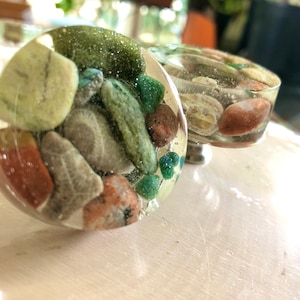 May include: Two round, clear resin knobs with embedded green, brown, and red stones. The knobs are approximately 5 cm in diameter.