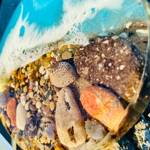 May include: A round resin coaster with a blue and white wave design and embedded stones and shells. The coaster is clear and shows the stones and shells through the resin.
