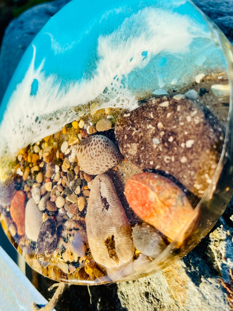 May include: A round resin coaster with a blue and white wave design and embedded stones and shells. The coaster is clear and shows the stones and shells through the resin.
