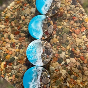 May include: Four round blue and white resin coasters with a beach scene design. The coasters are on a surface of small, colorful stones.