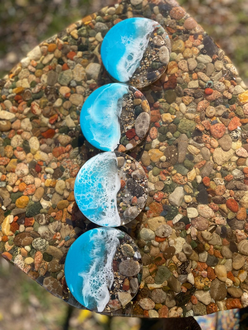 May include: Four round blue and white resin coasters with a beach scene design. The coasters are on a surface of small, colorful stones.