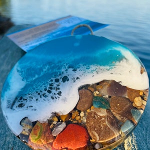 May include: A round resin art piece with a blue and white wave design and embedded stones. The wave is white and the water is a light blue color. The stones are various colors, including brown, red, and green.