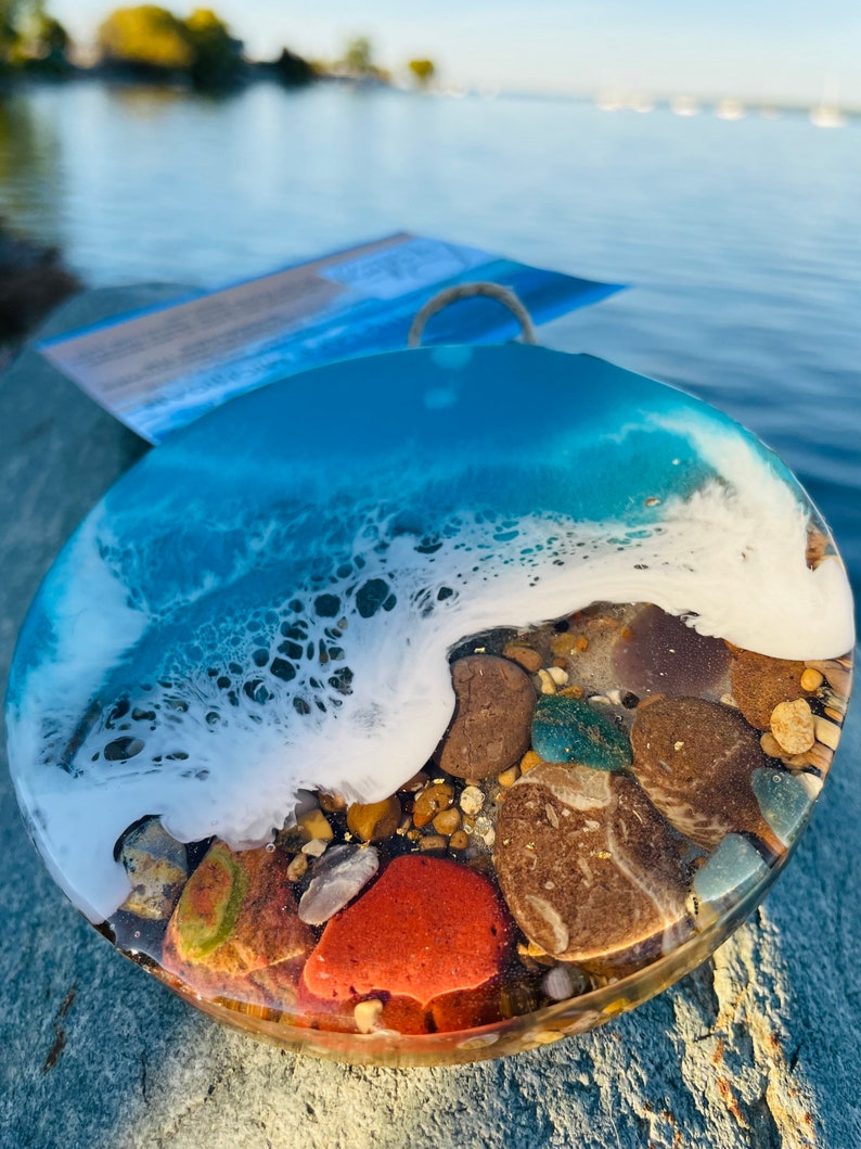 May include: A round resin art piece with a blue and white wave design and embedded stones. The wave is white and the water is a light blue color. The stones are various colors, including brown, red, and green.
