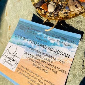 May include: A set of coasters featuring sand and stones collected from the shores of Lake Michigan. The coasters are inspired by the Great Lakes and are almost like a slice of Lake Michigan. The text on the card reads "Looking into Lake Michigan" and "Coasters inspired by the Great Lakes. Created with sand and stones collected along the shorelines."