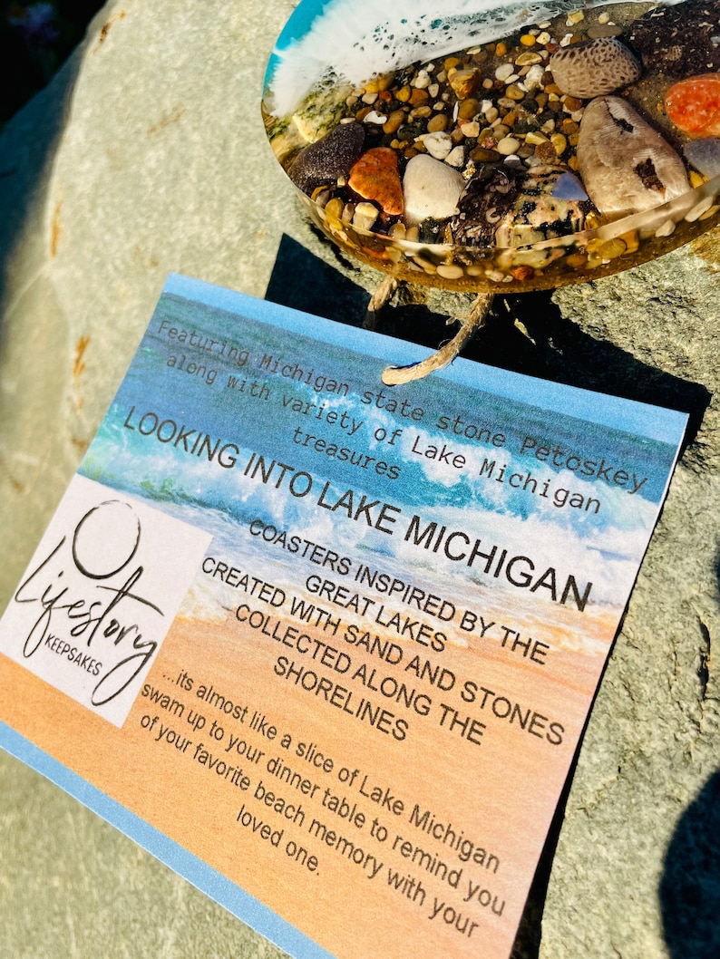 May include: A set of coasters featuring sand and stones collected from the shores of Lake Michigan. The coasters are inspired by the Great Lakes and are almost like a slice of Lake Michigan. The text on the card reads "Looking into Lake Michigan" and "Coasters inspired by the Great Lakes. Created with sand and stones collected along the shorelines."