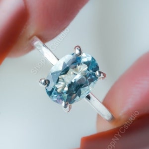 May include: A silver ring featuring a light blue oval gemstone. The gemstone is held in place by four prongs. The ring is held between two fingers with red nail polish.