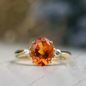 May include: A gold ring with an oval-shaped orange gemstone surrounded by two smaller clear gemstones.