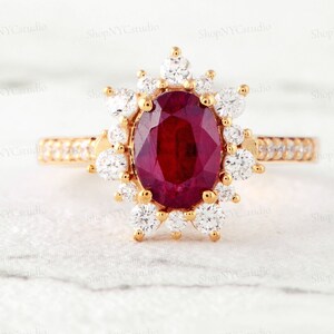 May include: A gold ring with a large oval ruby surrounded by a halo of smaller diamonds. The ring is set on a white background.