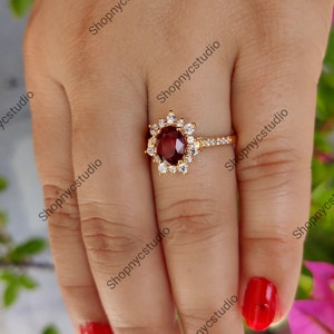 May include: A gold ring with a large, oval-shaped, red gemstone surrounded by smaller, white gemstones. The ring is on a finger with red nail polish.