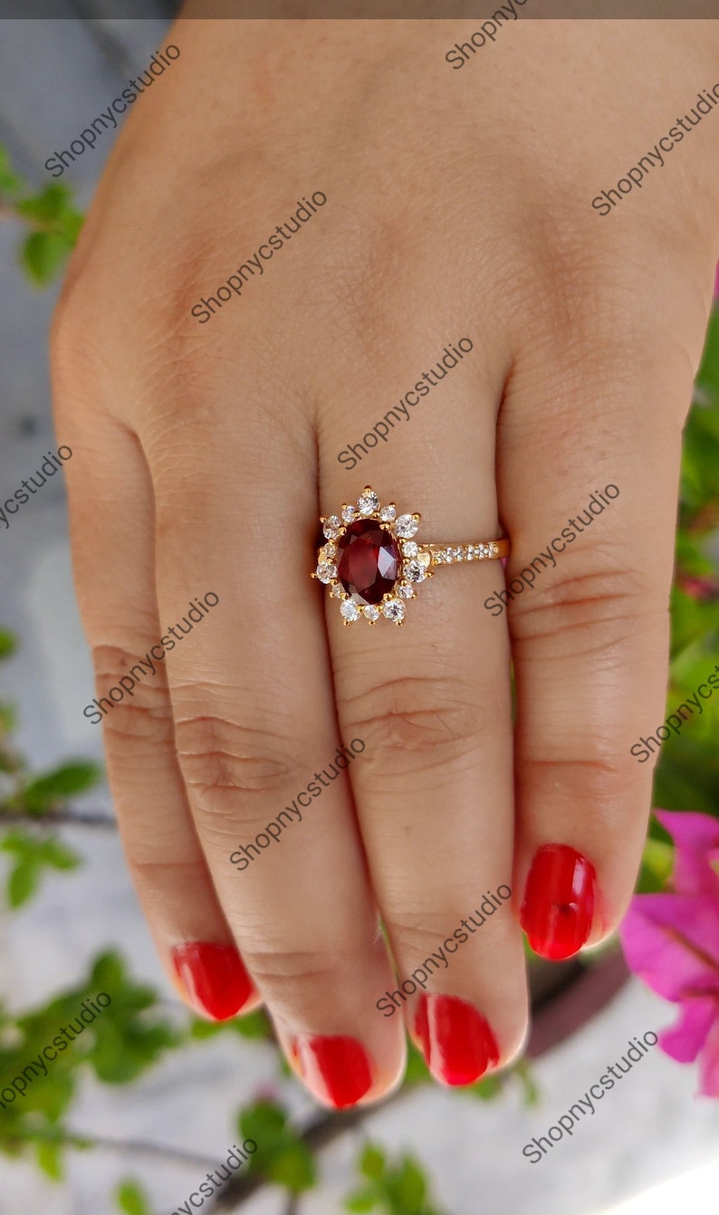 May include: A gold ring with a large, oval-shaped, red gemstone surrounded by smaller, white gemstones. The ring is on a finger with red nail polish.