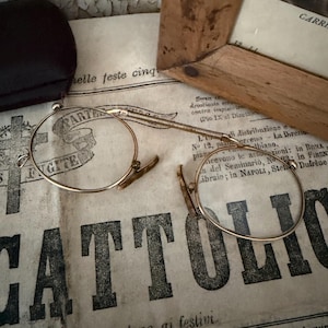 May include: A pair of gold-toned pince-nez spectacles featuring round wire rims connected by a flexible coiled metal bridge. The eyewear includes small, light-coloured nose pads attached to the inner edges of the circular frames.