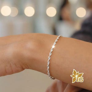 May include: A silver-toned rope chain bracelet with a twisted, textured design that wraps around the wrist. The metal surface features a reflective, polished finish that catches the light, creating a series of small, bright highlights along the links.