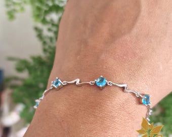 Peut inclure: Un bracelet à chaîne argentée orné de pierres rondes à facettes bleu vif, régulièrement espacées. Les maillons métalliques entre les pierres présentent une forme angulaire en zigzag, créant un motif continu autour du poignet.