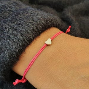 Peut inclure: Un bracelet en cordon rose fin doté d'une petite breloque métallique en forme de cœur lisse centrée sur le fil. Le bracelet est fixé par des extrémités nouées réglables, créant un design simple et délicat qui repose sur la peau.