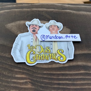 May include: A sticker featuring two men in white suits and hats, with the text "Los Dos Carnales" in yellow and black lettering. The sticker also includes the social media handle "@Mendoza.Arte".