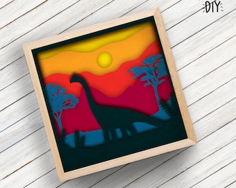 Pode incluir: Uma obra de arte em papel emoldurada e em camadas com a silhueta de um dinossauro contra um pôr do sol. A obra usa tons de laranja, vermelho e azul para retratar uma cena pré-histórica. A moldura é de madeira clara, com o texto "KOOTSIE DIY".