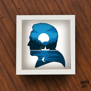 May include: A framed paper art piece showing a man's head silhouette. The interior reveals a layered blue scene of a lake, a fisherman in a boat, and a rising sun. The white frame contrasts with the wooden background.